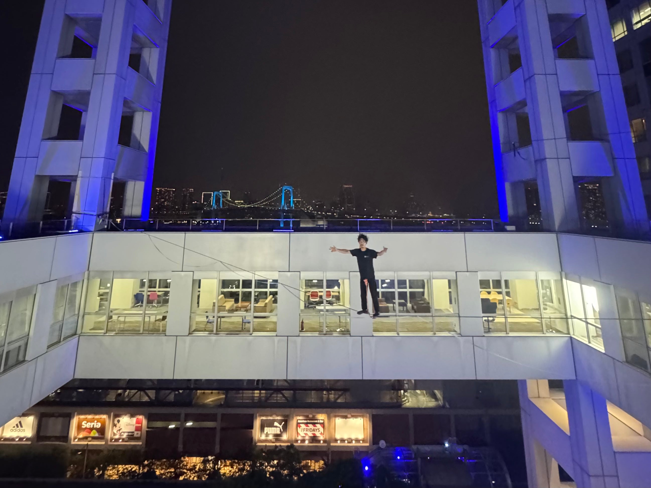 フジテレビ『ENGEI 8』ハイライン企画 お台場フジテレビ本社ビルの空中に張られたハイラインの上に立つ劇団ひとりさん 背景にレインボーブリッジの夜景 Slackline Researchが設置・安全監修